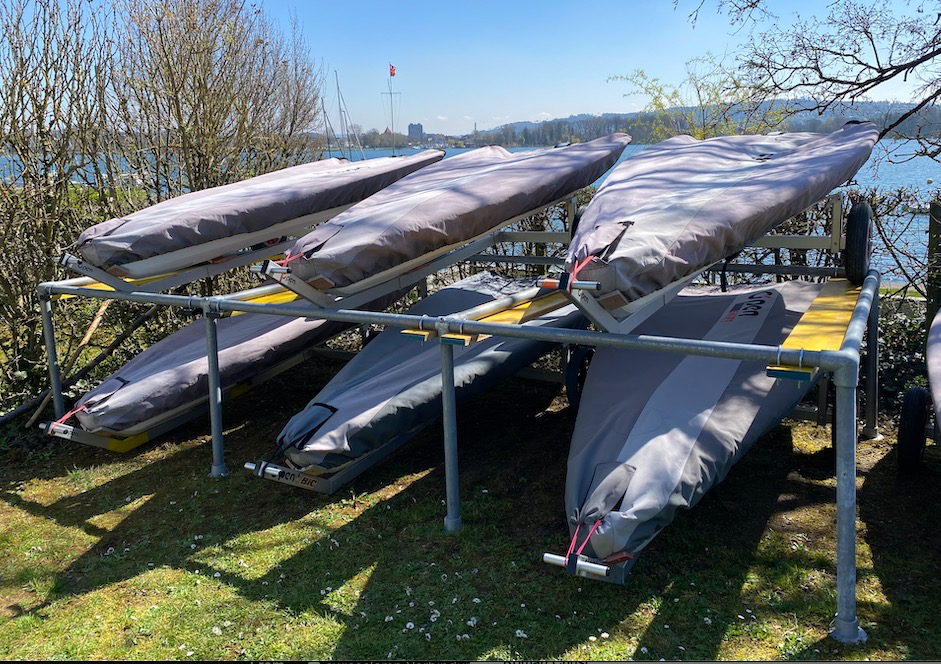 Mehrere kleine Segelboote, mit grauen Planen abgedeckt, stehen auf einem Metallgestell am Seeufer. Vorn grünes Gras, dahinter blaues Wasser mit weiteren Booten und Gebäuden am Horizont unter klarem Himmel. Lange Schatten.