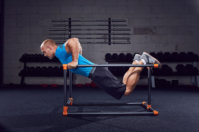 Ein muskulöser Mann in blauem Tanktop und schwarzen Shorts macht eine fortgeschrittene Bodyweight-Übung am Dip-Ständer im Fitnessstudio. Beine angewinkelt auf dem Holm, Oberkörper parallel zum Boden, trainiert Brust und Trizeps.