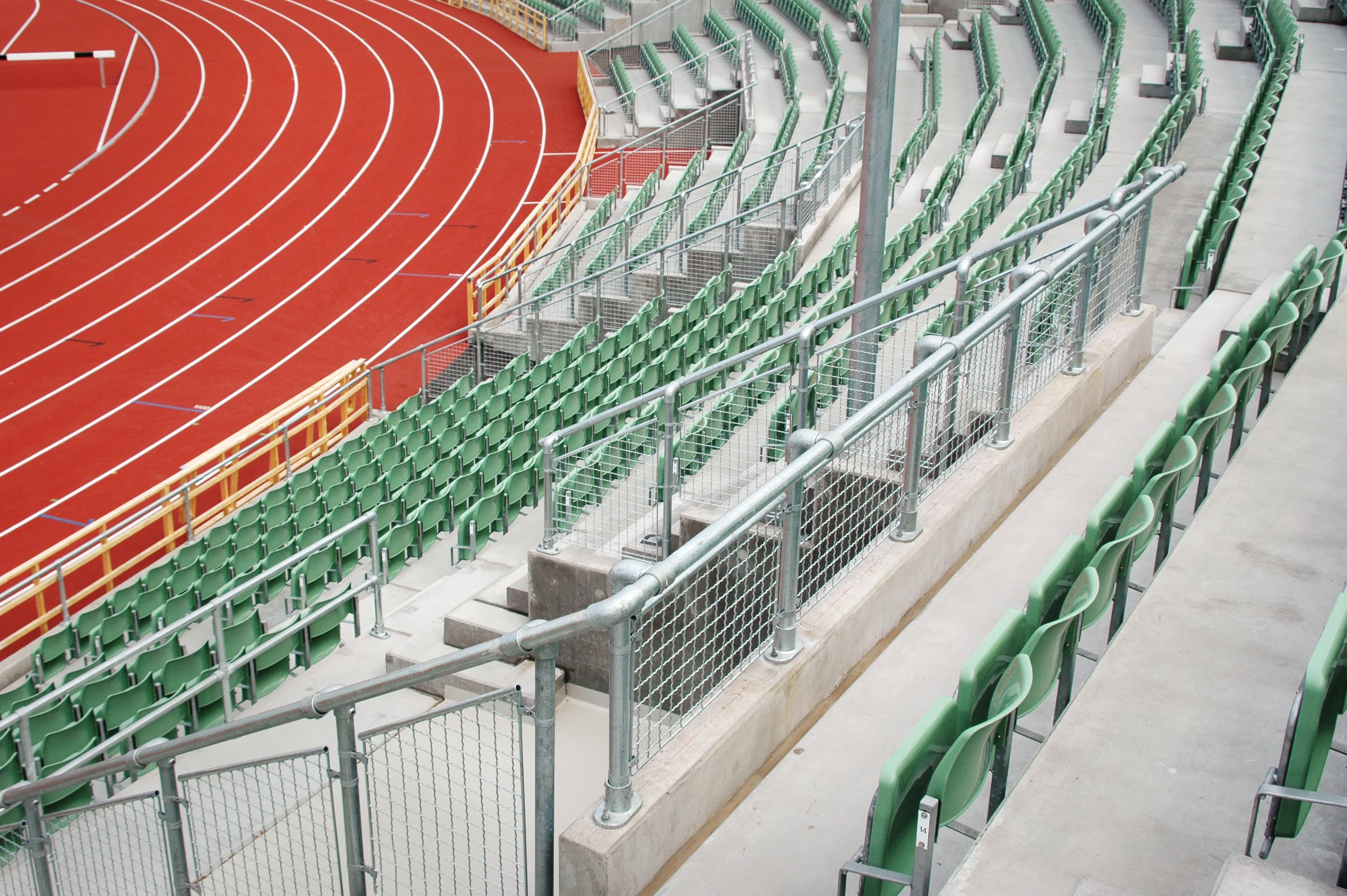 Eine leere rote Laufbahn mit weißen Spurlinien liegt links und biegt in der Kurve nach rechts. Rechts und im Hintergrund sind leere grüne Stadionsitze auf Betonstufen, getrennt durch Metallgeländer mit Gittern.